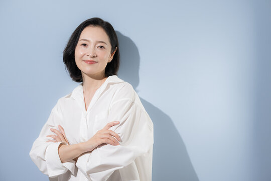 [水色背景]自信を持って腕を組む50代の日本人女性、管理職や経営者、専門家、キャリア、信頼を表現するオフィスカジュアルのビジネスイメージ