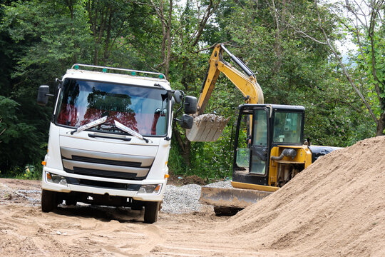 Construction truck and excavator near sand pile at wooded outdoor worksite, showing heavy equipment during road preparation and earthmoving activity
