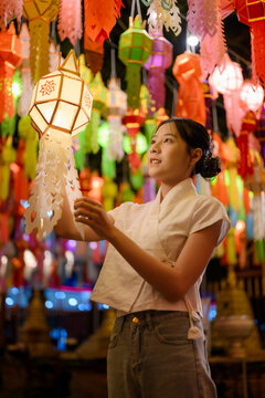 Young Asian woman holding traditional glowing paper Lanna lantern during Yi Peng lantern festival night under colorful hanging lights