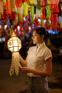 Young Asian woman holding traditional glowing paper Lanna lantern during Yi Peng lantern festival night under colorful hanging lights