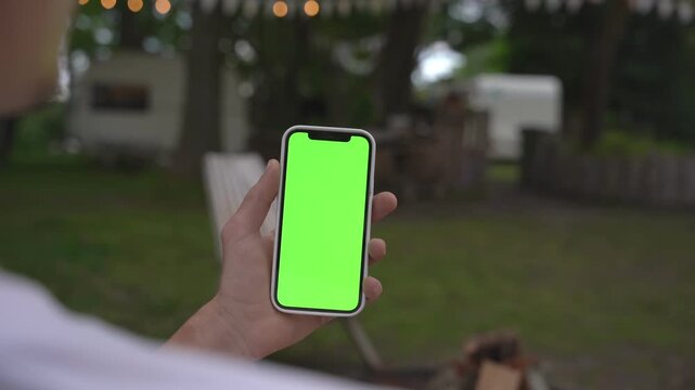 Hand holds phone with green screen outdoors at dusk. Background shows blurred backyard with trees and soft lights. Person wears white shirt, facing away from camera. Footage ideal for app demos