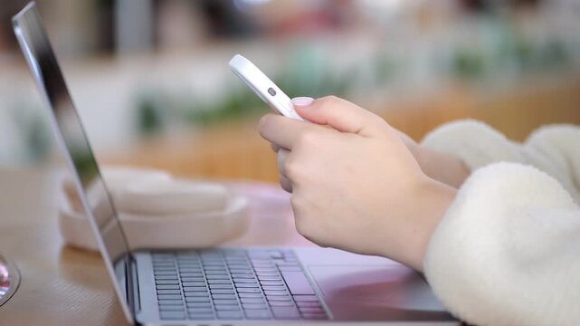 Close-up hands holding phone beside laptop on wooden table. Soft, warm lighting in background. Person wears cozy white sweater. Scene shows modern digital multitasking