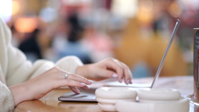 Close-up of hands typing on a laptop in a cafe. White ceramic mug sits beside the device. Soft bokeh background shows indistinct people. Warm wooden table surface under the hands