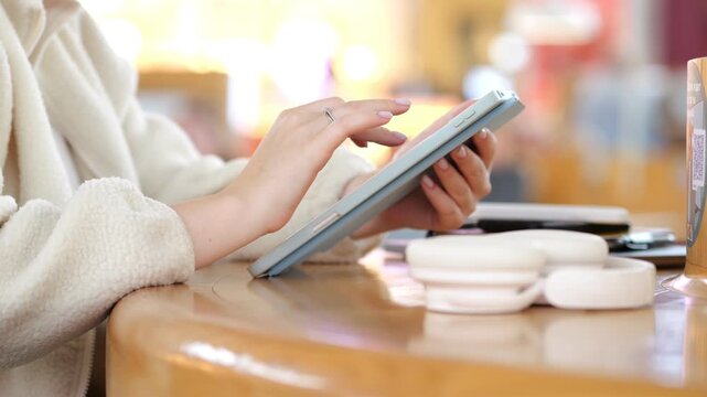 Hands use tablet on wooden table. White sweater, blurred background. Coffee shop setting. Soft lighting, warm tones. Focus on interaction with digital device