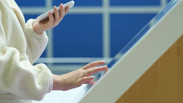 Person uses smartphone and touches transparent screen. Blue windows behind. Light wood frame visible. Hands in cream fleece sleeves. Modern interactive surface shown
