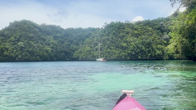 Kayaking through turquoise waters of remote tropical lagoon with white sailboat anchored in distance.