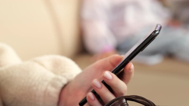 Close-up of a person's hand holding a phone. The hand has a ring and light-colored nails. Background is softly blurred with indoor lighting. Child is visible out of focus in the distance
