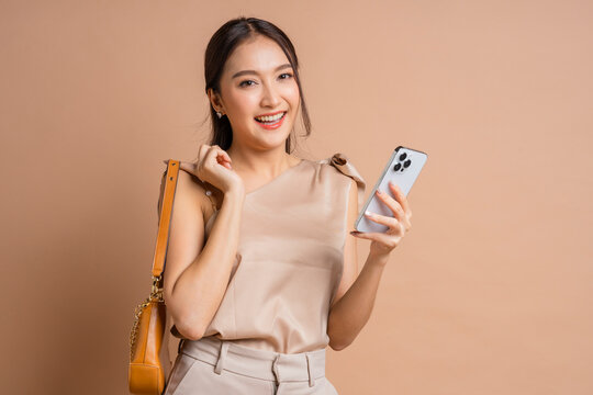 Happy smiling Asian woman holding handbag and smartphone in beige studio, demonstrating fashionable shopping lifestyle, modern mobile banking promotion, consumer satisfaction, and retail elegance.