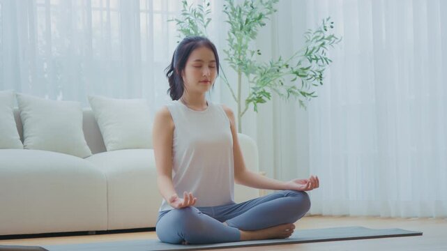 Happy young Asian woman practicing yoga and meditation at home sitting on floor in living room in lotus position and relaxing with closed eyes. Mindful meditation and wellbeing concept