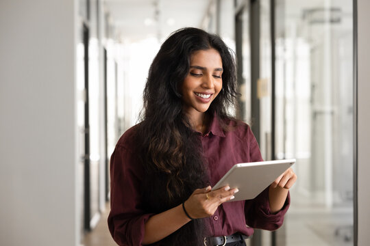 South Asian businesswoman holding digital tablet in modern office hallway, reviewing project details, using application for business communication and corporate tasks management, reading work emails