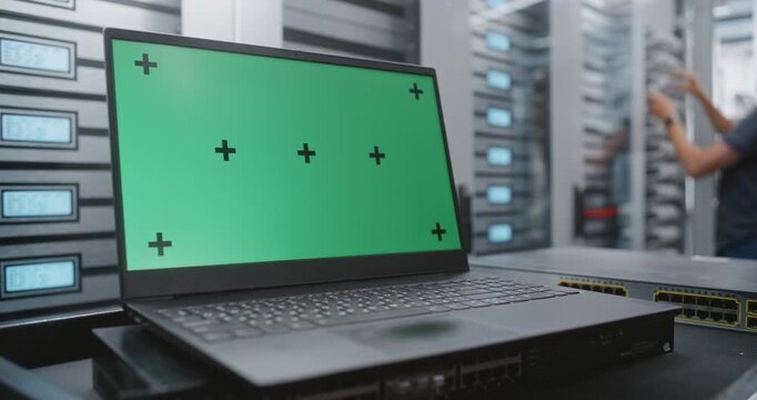 Laptop With Chroma Key Display Stands Beside Server Cabinets in Professional Data Center, Creating Adaptable Footage for Replacing Screen Content With Dashboards, Applications, Digital Graphics.