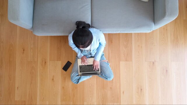 Top view of young Asian woman sitting on the floor and working on computer laptop with mobile smart phone in living room at home