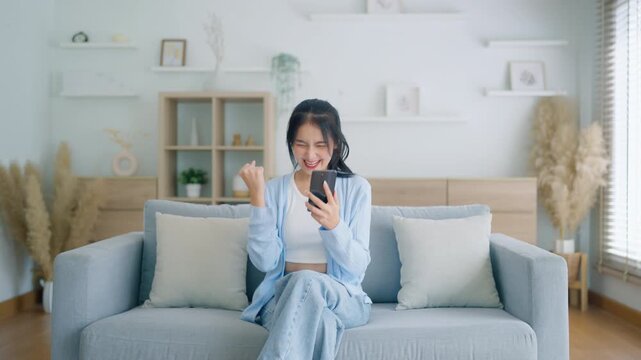 Smiling pretty young Asian woman sitting on cozy couch, using funny mobile apps in living room. Woman at Home, Doing Online Shopping, Messaging Friends, Posting on Social Media