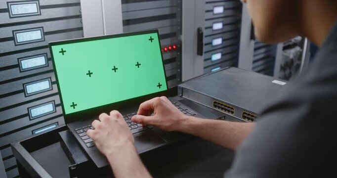 Male IT Engineer Works on Laptop With Green Screen Display Beside Server Racks, Providing Customizable Screen Replacement Footage For Cybersecurity, Cloud Computing and Enterprise Technology Concepts.