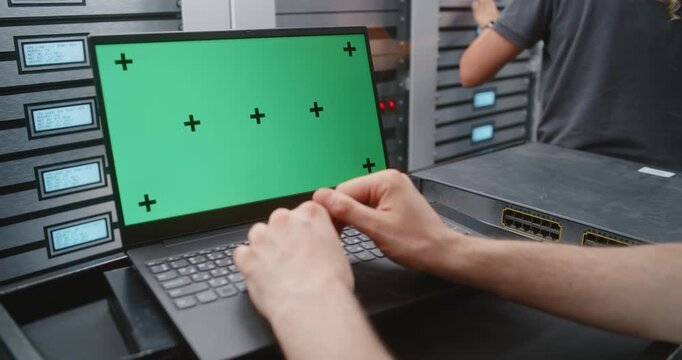 Male System Administrator Uses Laptop With Green Screen Beside Modern Server Cabinets, Creating Flexible Footage For Replacing Display Content With Monitoring Dashboards, Software Interfaces and IT