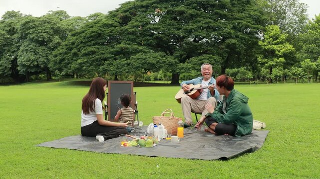 GrandFather playing acoustic guitar for child during picnic. Asian family enjoy outdoor picnic with food and drinks in park sunny day, outdoor activitie with daughter father, mother grandfather