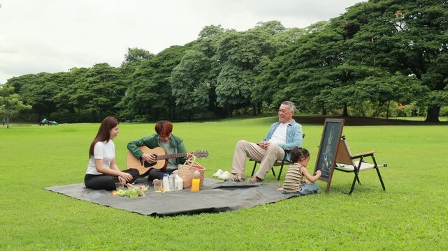 GrandFather playing acoustic guitar for child during picnic. Asian family enjoy outdoor picnic with food and drinks in park sunny day, outdoor activitie with daughter father, mother grandfather