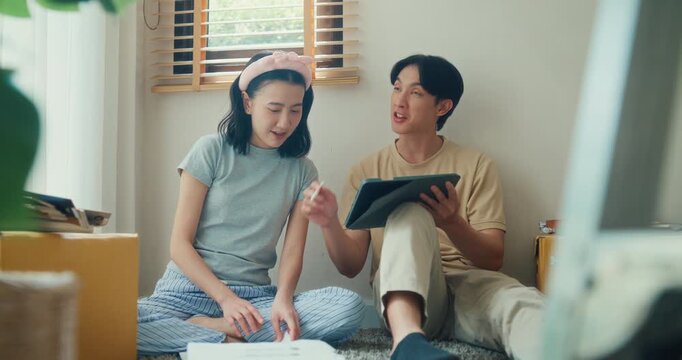 Young Asian couple plans move at home while browsing furniture online on tablet beside packed boxes. Relocation planning, new home setup, online shopping, household design and unpacking concept.