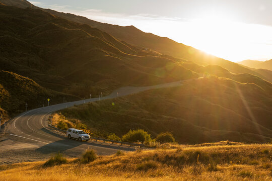 Vehicle on a winding mountain road at golden sunset above tussock hills, New Zealand