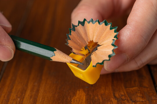 Close-up of human hands sharpening a green wooden pencil with a yellow manual sharpener, showing wooden shavings on a wooden table.