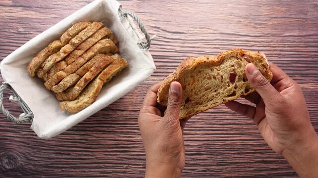 Top-down view of a person&rsquo;s hands slowly breaking a slice of freshly baked sourdough bread in two pieces, with a bread basket full of sliced bread in the background