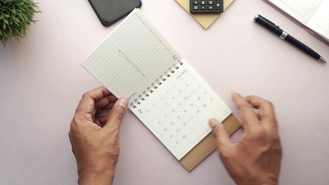A male hand flipping through the pages of a 2023 desk calendar showing a close-up, top-down view of the monthly schedule with a focus on future planning, deadlines, and time management