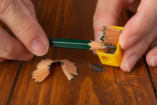 Close-up of human hands sharpening a green wooden pencil with a yellow manual sharpener, showing wooden shavings on a wooden table.