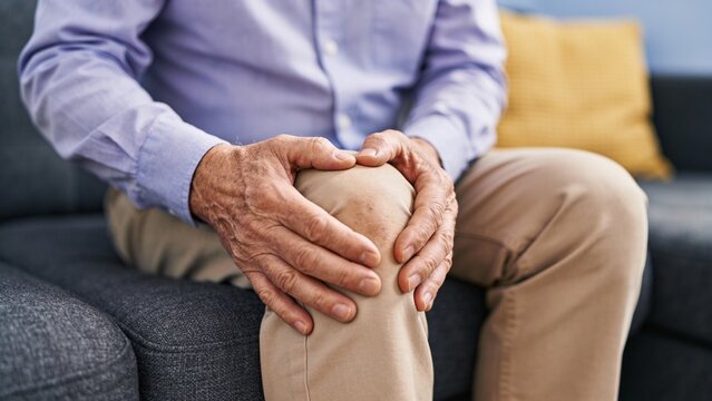 Senior man holding painful knee while sitting on a sofa indoors