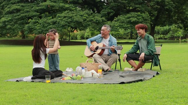 GrandFather playing acoustic guitar for child during picnic. Asian family enjoy outdoor picnic with food and drinks in park sunny day, outdoor activitie with daughter father, mother grandfather