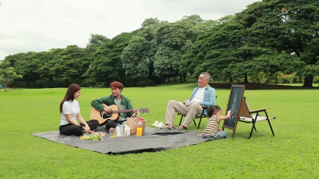 GrandFather playing acoustic guitar for child during picnic. Asian family enjoy outdoor picnic with food and drinks in park sunny day, outdoor activitie with daughter father, mother grandfather