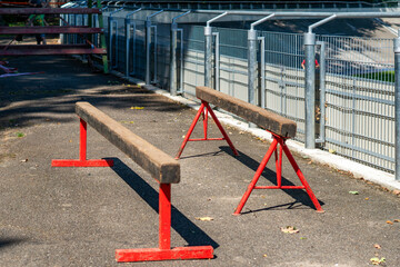 View of two wooden balance beams mounted on red metal stands set on an asphalt surface next to a metal fence and protective railings on a sunny day.    © Adam