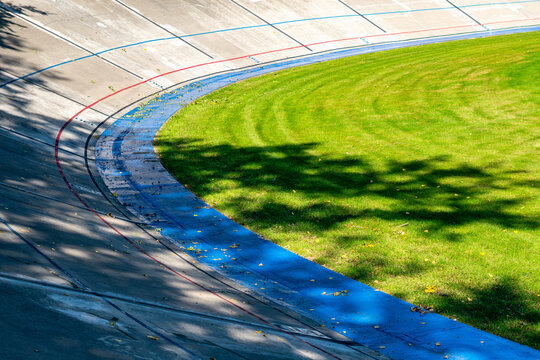 High angle view of a concrete cycling velodrome track curve featuring painted measurement lines, a blue inner band, and a green grass field inside the oval in sunny daylight with tree shadows.   
