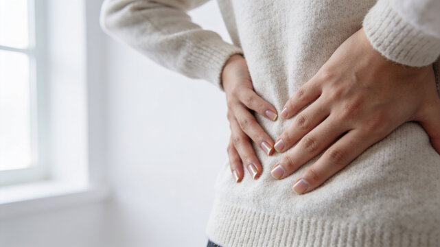 Person in a cream knit sweater pressing both hands to the lower back, showing back pain, lumbago and spine strain. Backache, sciatica and posture health concept in soft light.