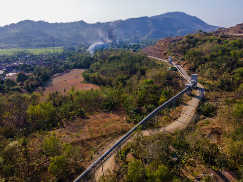 Aerial View of Covered Conveyor Belt System Checkpoint Buildings and Service Road Connecting an Industrial Cement Plant and Large Storage Dome Across Hilly Terrain in Indonesia. 