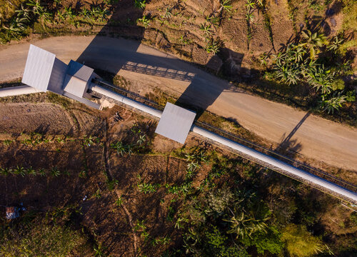 Aerial Scenery of Covered Conveyor Belt System with Checkpoint Buildings and a Service Road Crossing Hilly Terrain in Indonesia. Industrial Infrastructure Extends Across the Green Hillside. 