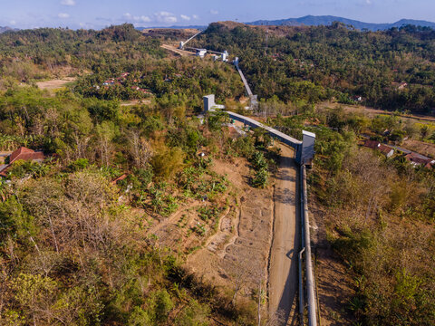 Aerial View of a Covered Conveyor Belt System with Checkpoint Buildings and a Service Road Crossing Hilly Terrain Near a Residential Area in Indonesia