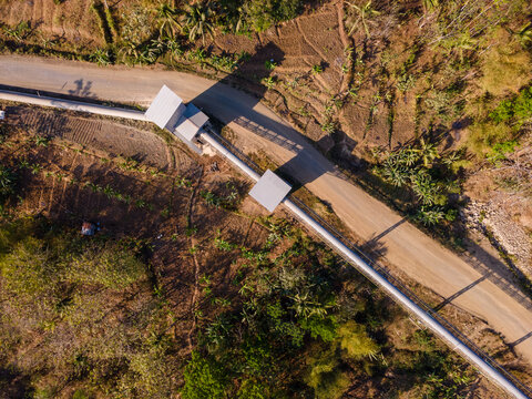 Aerial View of a Covered Conveyor Belt System with Checkpoint Buildings and a Service Road Crossing Hilly Terrain Near a Residential Area in Indonesia
