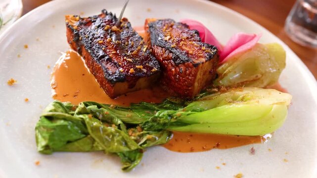 Close up video of hands using fork and knife to cut into a perfectly cooked pork belly dish, showcasing the juicy meat and crispy skin on a white plate with greens and sauce.