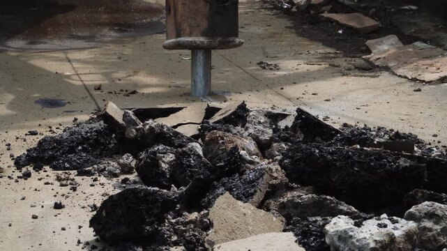 Static tripod footage shows a hydraulic rock breaker attachment striking and breaking an asphalt road surface, with fractured pavement, rubble, dust, and debris visible during road repair work.