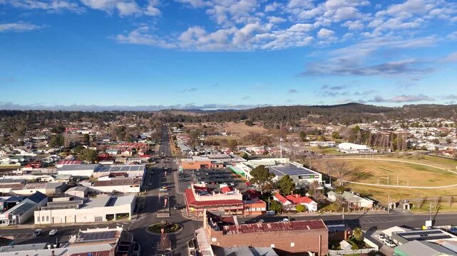 Aerial view of a rural town with a main road, roundabouts, and local businesses under a vibrant blue sky with scattered clouds.