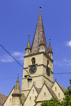 The Sibiu Lutheran Cathedral's tower with its distinctive colorful tiled roof and clock faces against a clear blue sky.