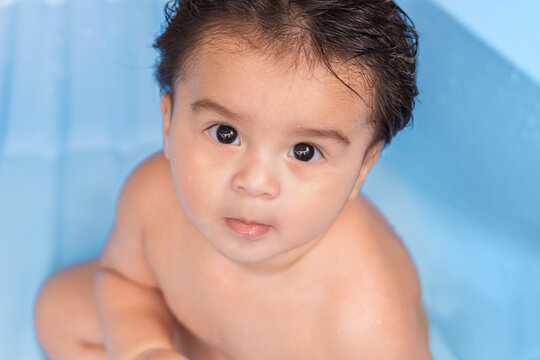 Portrait of a baby with wet hair looking at the camera during bath time against a blue background. Infant hygiene, care and everyday routine concept.