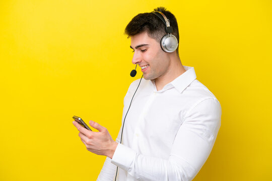 Telemarketer caucasian man working with a headset isolated on yellow background keeping a conversation with the mobile phone with someone