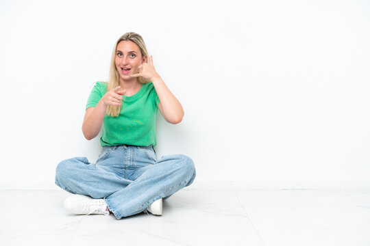 Young caucasian woman sitting on the floor isolated on white background making phone gesture and pointing front