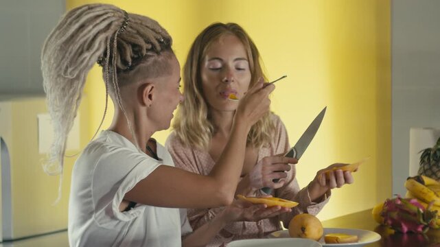 Two women cutting mango at kitchen counter with knife, sliced fruit and peel on plate, warm daylight kitchen background