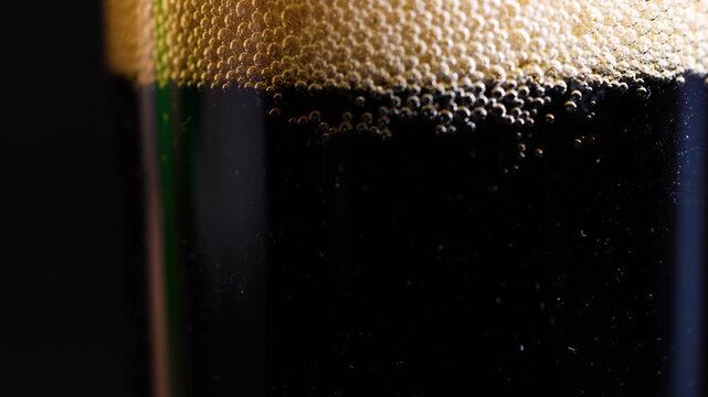 Close up motion of dark soda bubbles settling in transparent glass creating dynamic frothy texture on black background.