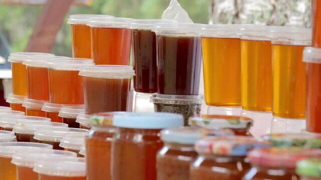 Stacked honey and syrup containers displayed on market table with warm daylight reflections, with honey, syrup, jar, container detail, showing lid, amber, golden, brown context