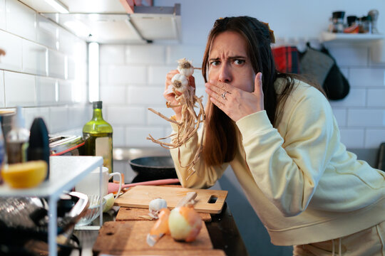 
Woman Covering Mouth While Holding Smelly Fresh Garlic Bulbs. Shocked female reacting to strong odor while holding organic garlic in kitchen