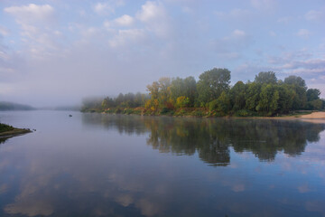 Obraz premium A peaceful river flows calmly under a cloudy sky, reflecting the green trees lined along the misty distant shoreline
