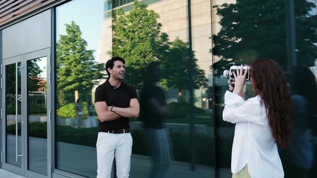 Woman filming a handsome man posing in front of a modern glass building with a retro camera